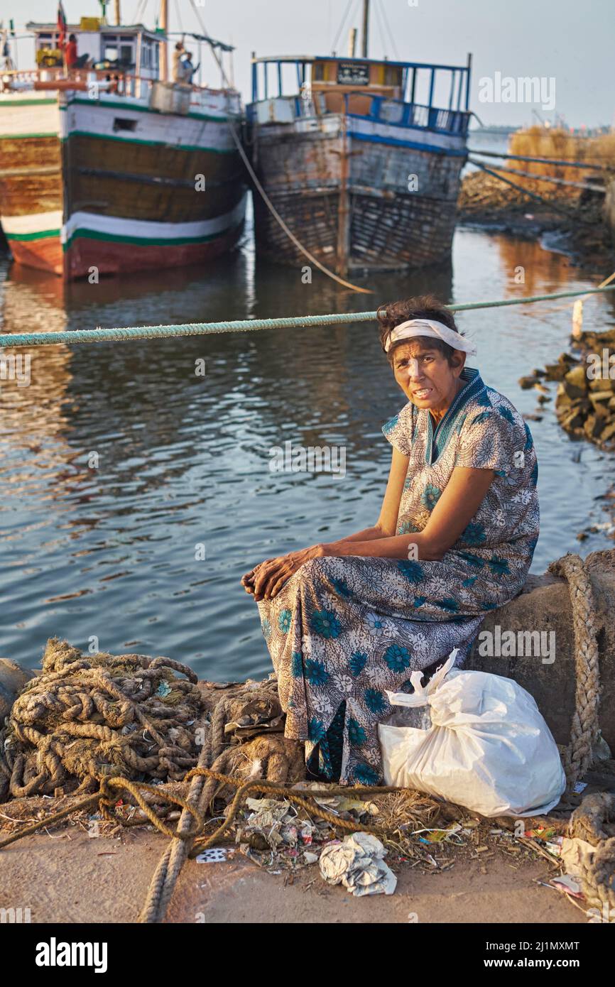 A middle-aged Indian woman sits alone in a far-flung corner of the Old Port in Mangalore (Mangaluru), Karnataka, South India, fishing boats in the b/g Stock Photo