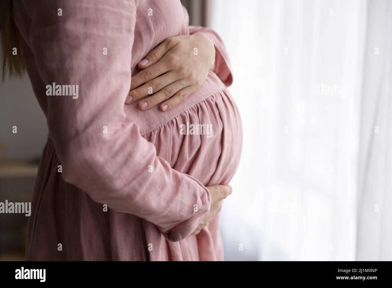 Dreamy young pregnant woman standing near window. Stock Photo