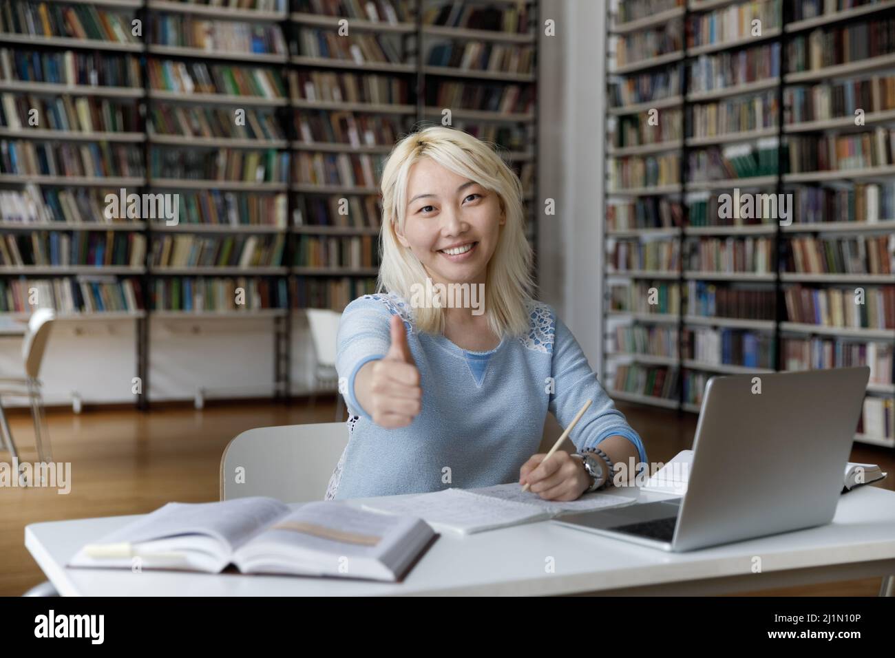 Asian student shows thumbs up seated at table in library Stock Photo ...