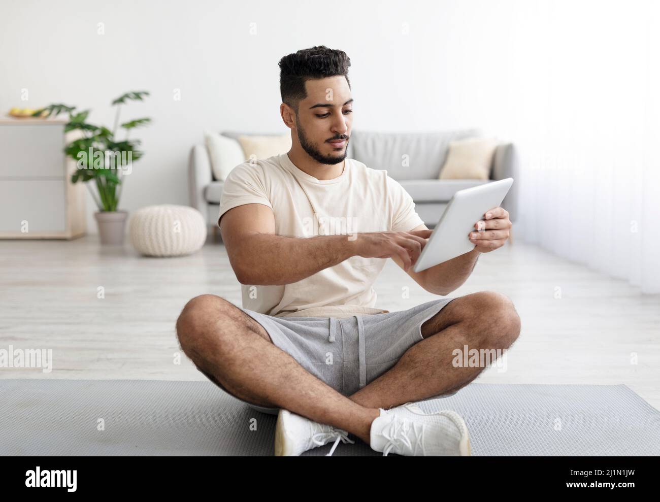 Young Arab guy using tablet computer while sitting cross legged on yoga mat at home Stock Photo