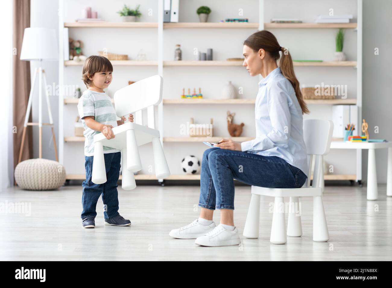 Furious emotional little boy grabbing chair in anger, shocked woman psychologist trying to work with difficult patient Stock Photo