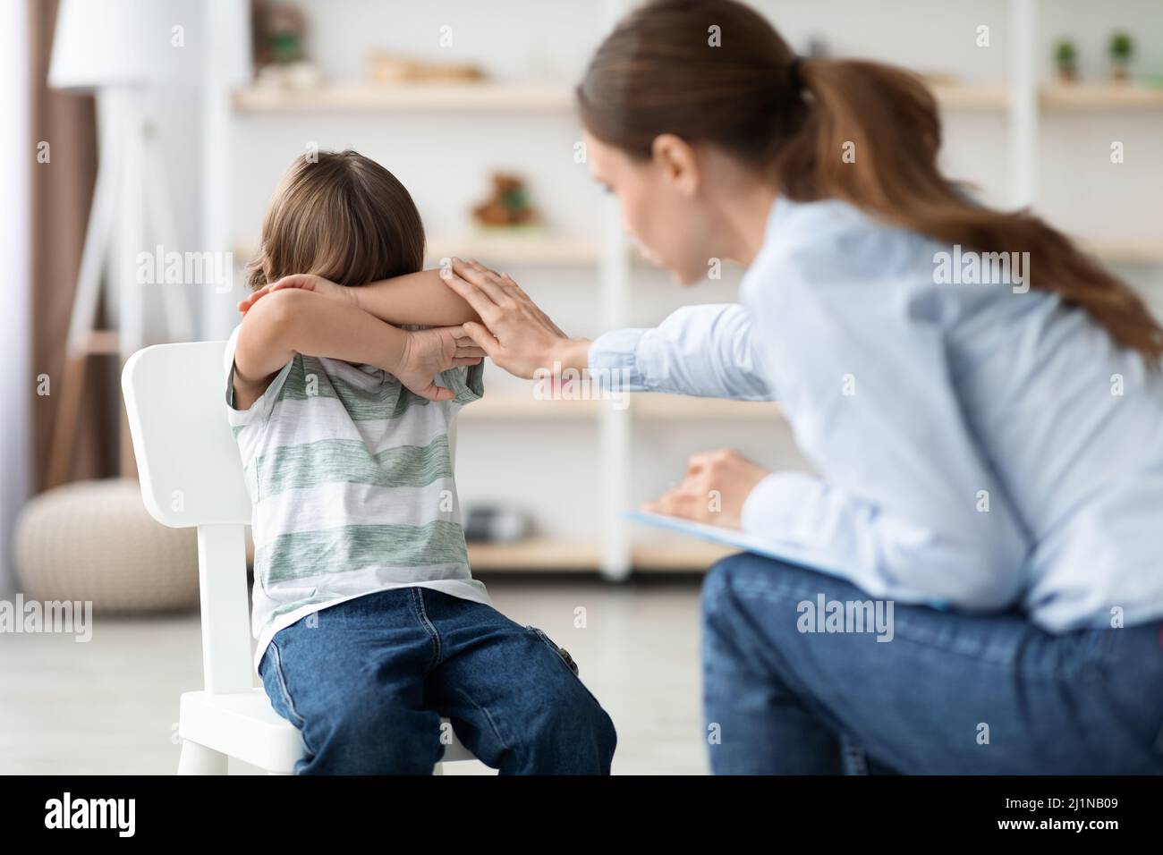 Kids communication blocks. Little scared boy hiding face behind hands, ignoring caring woman psychologist Stock Photo