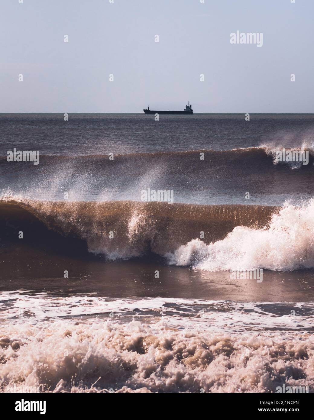 Atlantic ocean waves breaking near the windy coast in Portugal and a small coastal tanker or tank ship far away in the horizon. Vertical composition Stock Photo