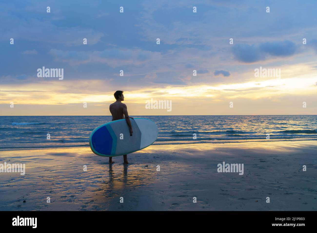 Asian man holding a sup board and paddle and walking on the beach. In the background, the ocean and the sunset. Back view. Summer surfing. Stock Photo
