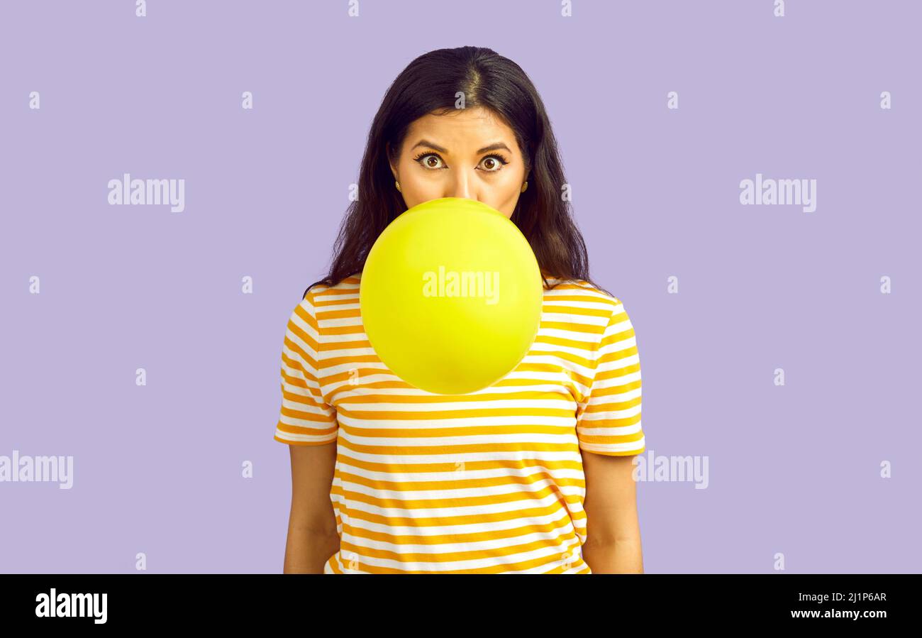 Woman isolated on lilac background blowing up yellow balloon with funny face expression Stock Photo