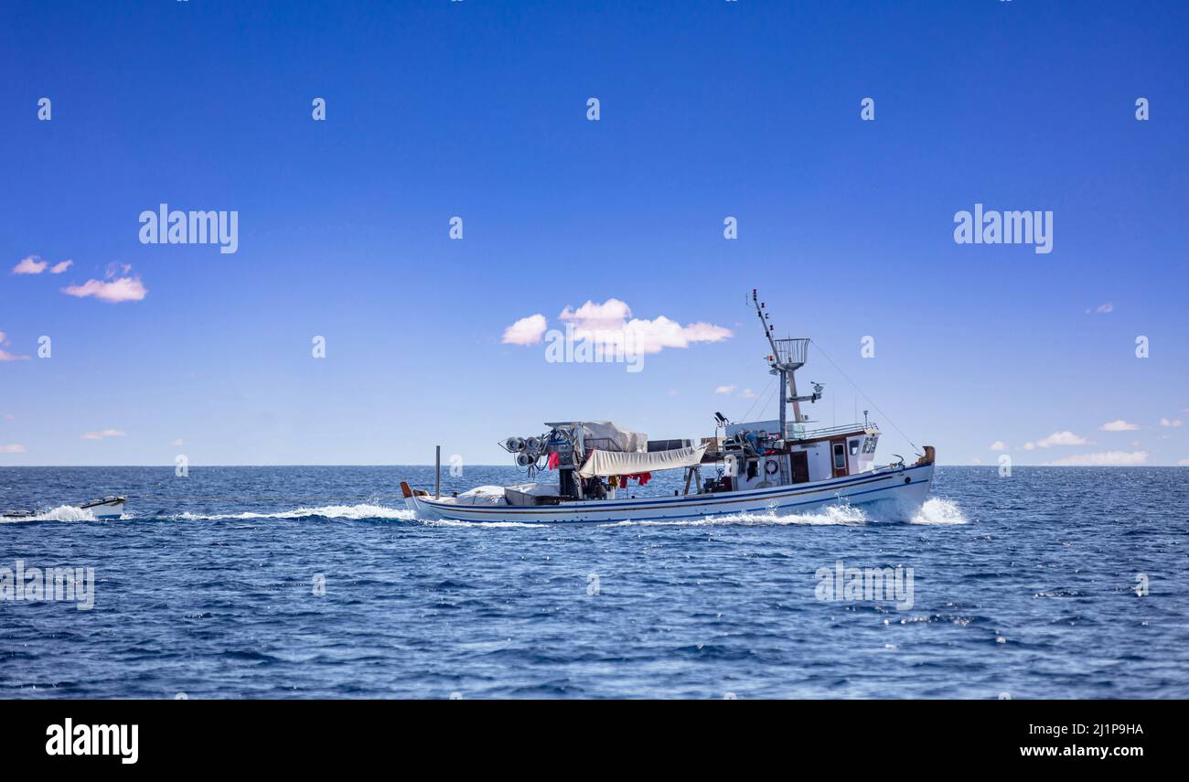 Fishing boat return. Trawler loaded with fish sails in Aegean rippled sea, a small vessel tied with rope follows, blue sky background. Cyclades Greece Stock Photo