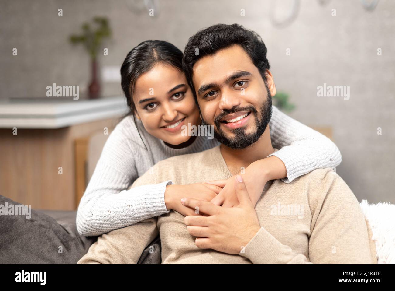 Portrait of lovely couple at home. Handsome man sitting on the comfortable couch, beautiful woman hugging his from the back. Spouses spend time together Stock Photo