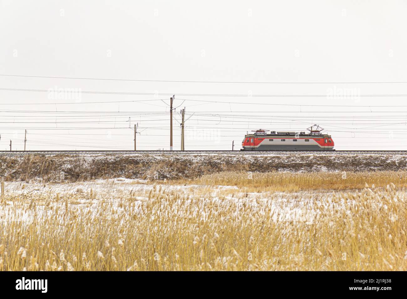 A lone electric train without wagons moves along the railway past yellow grass against the backdrop of white snow. Stock Photo