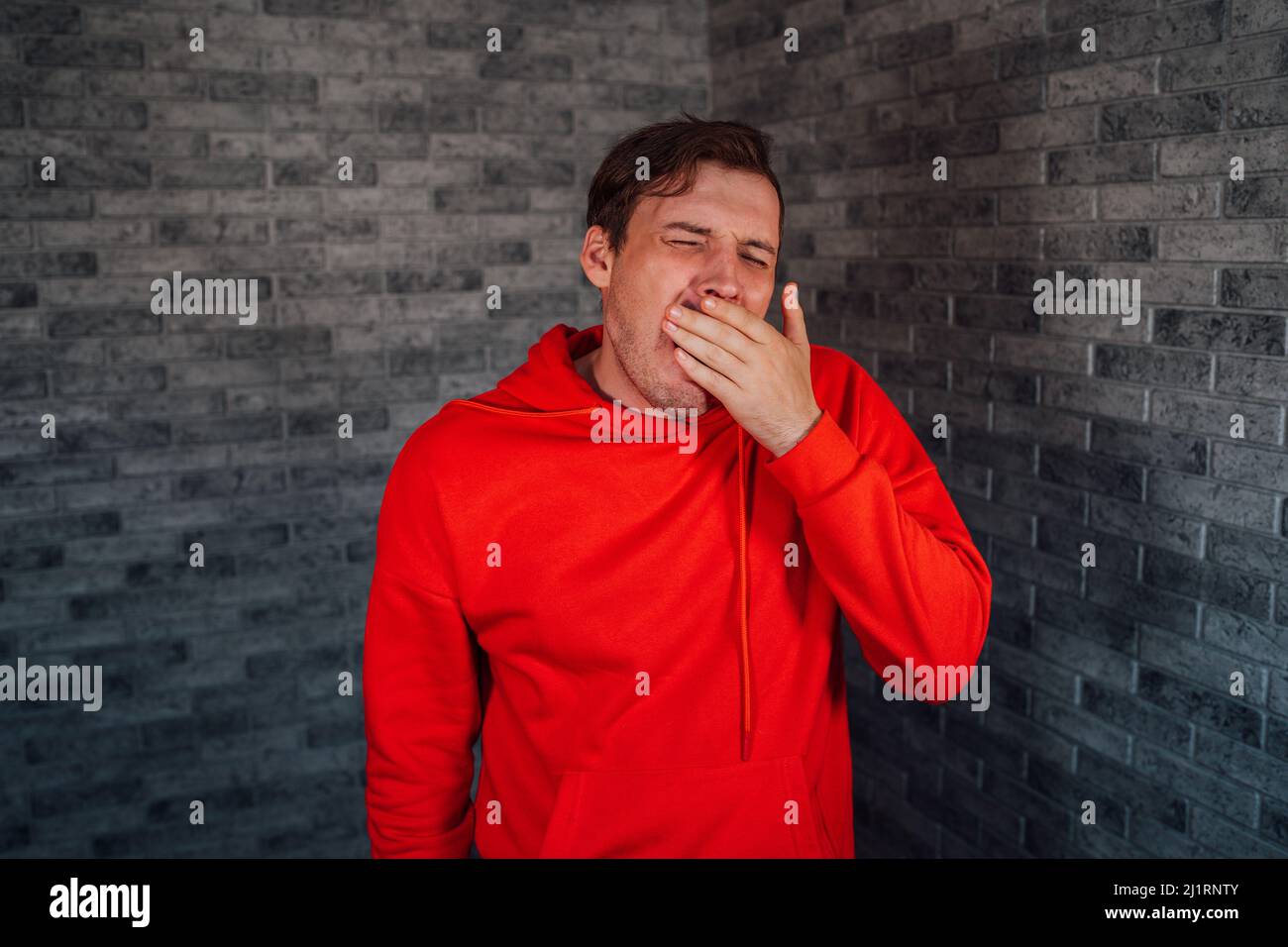 man in a red hoodie yawns from fatigue on a brick background. Stock Photo