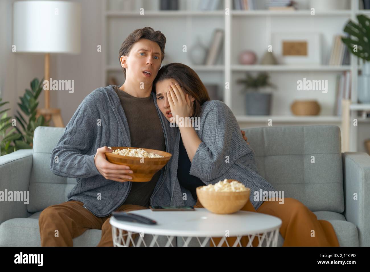 Shocked family with popcorn sitting on sofa and watching TV at home. Couple spending time together. Stock Photo