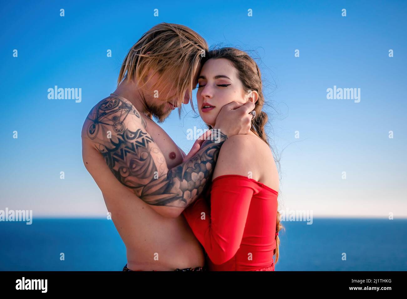 Portrait of tattooed man and sensual young brunette woman in red dress, happily dancing outdoors isolated on blurry sea background with natural bokeh Stock Photo