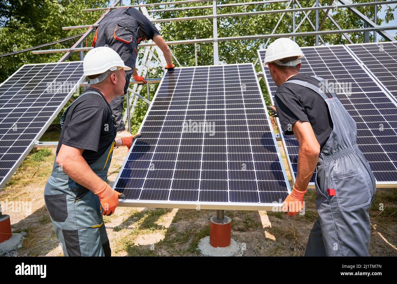 Workers building photovoltaic solar panel system outdoors. Men engineers placing solar module on metal rails, wearing construction helmets and work gloves. Renewable and ecological energy. Stock Photo