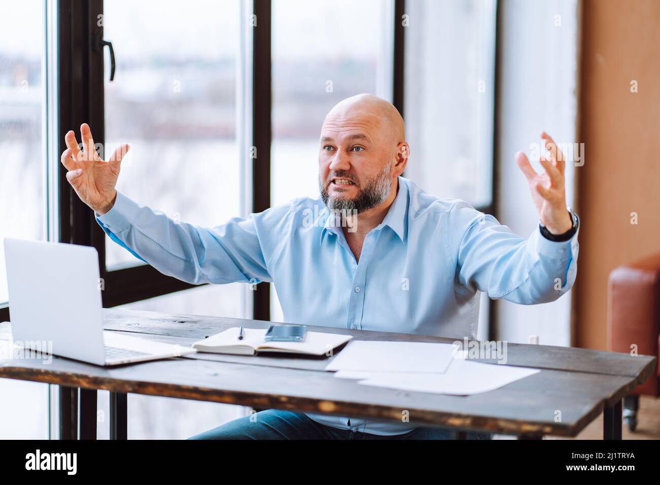 Portrait of angry annoyed middle-aged bald man wearing blue shirt sitting at wooden desk near document laptop in office. Stock Photo