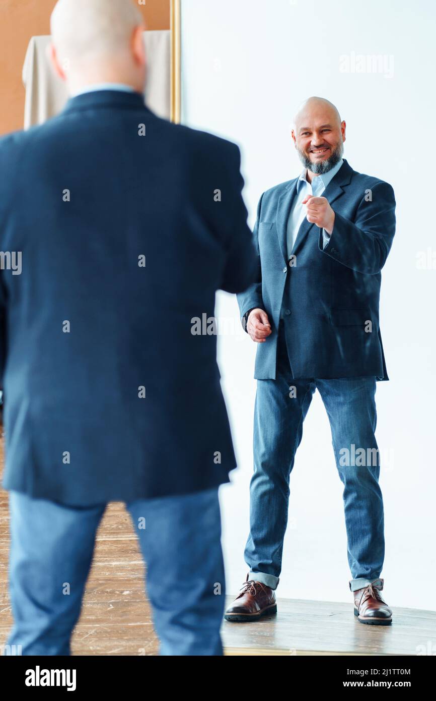 Portrait of happy middle-aged bald man with gray beard wearing blue suit jacket, pointing finger at mirror reflection. Stock Photo