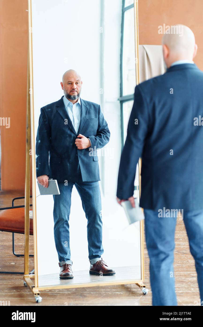Portrait of serious middle-aged bald man with gray beard wearing blue suit jacket, holding notebook, looking at mirror. Stock Photo