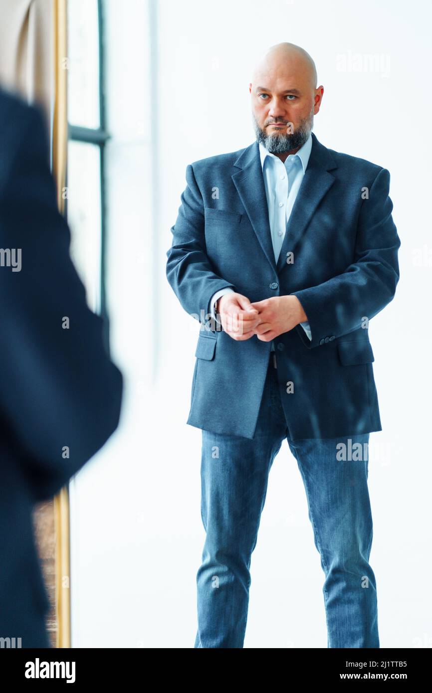 Portrait of serious middle-aged bald man with gray beard wearing blue suit jacket, looking gravely at mirror reflection. Stock Photo
