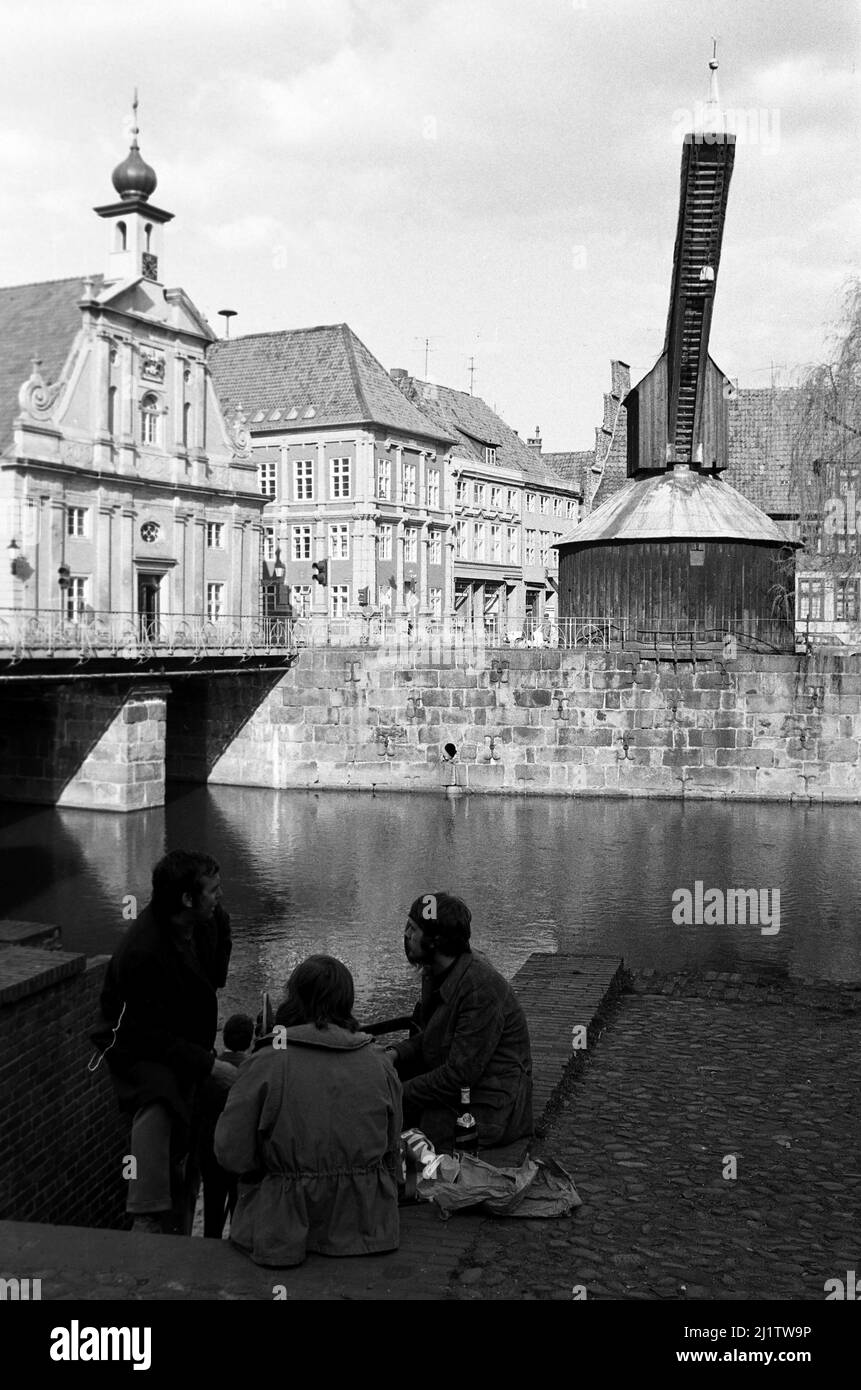 Junge Menschen sitzen am Ufer der Ilmenau mit Blick auf den Alten Kran im Lüneburger Hafen, 1970. Young people sitting on the shore of Ilmenau river with view of the Old Crane at the Lüneburg Port, 1970. Stock Photo