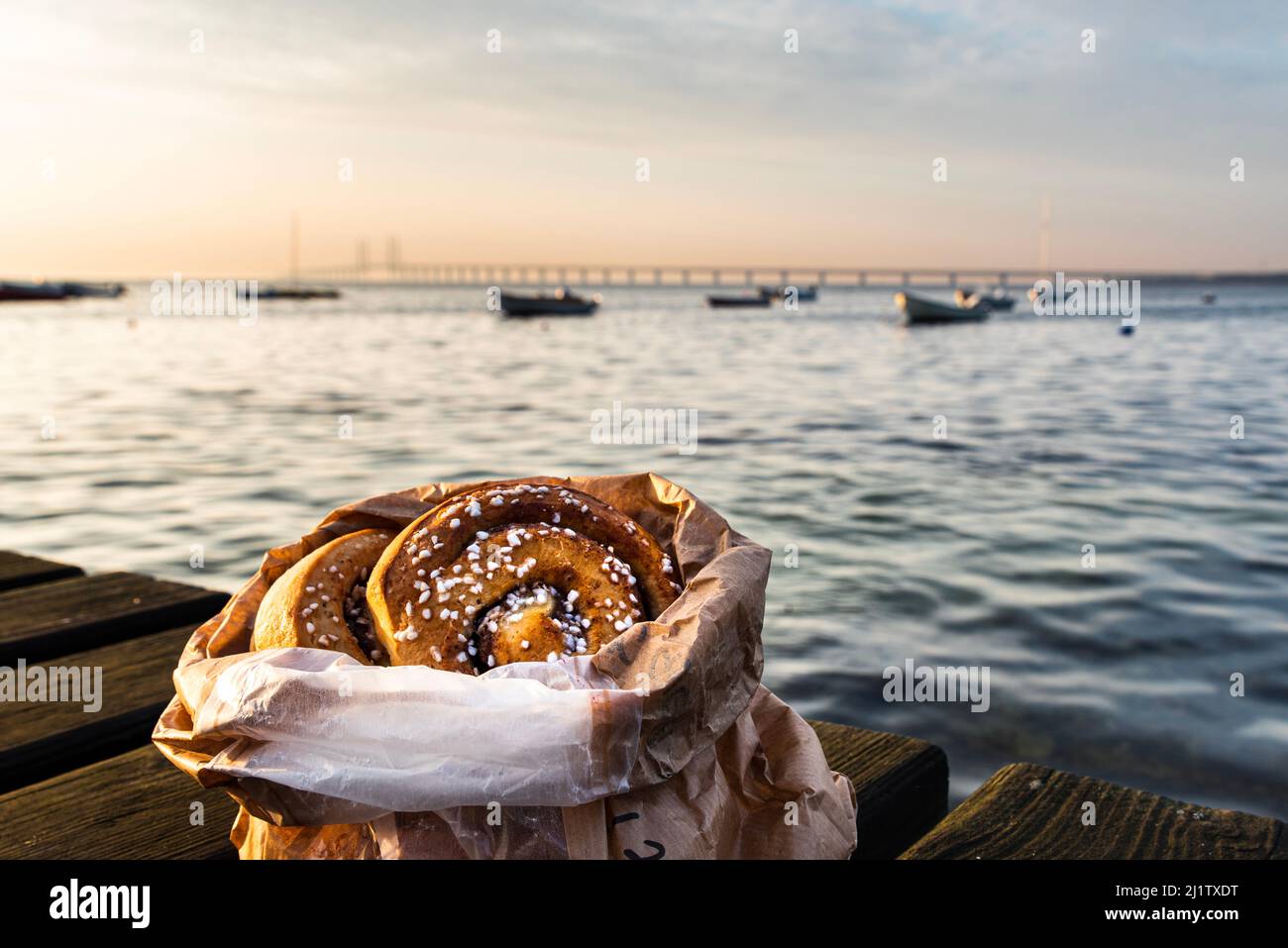 Cinnamon roll on a wooden boardwalk by the ocean at dusk with the Oresund Bridge. Cosy Swedish fika by the sea at sunset, typical Scandinavian pastry Stock Photo
