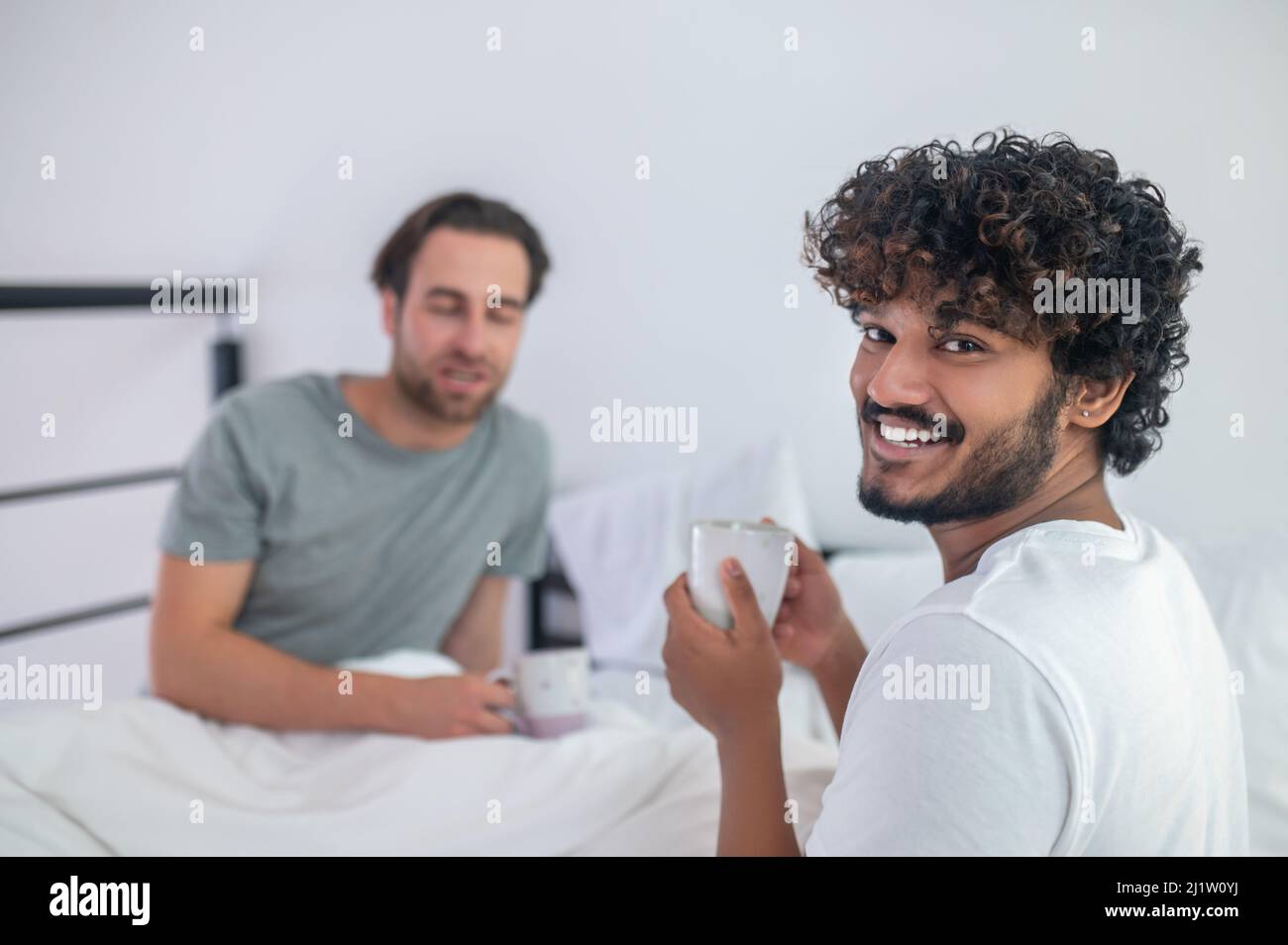 Man drinking coffee with his sleepy boyfriend in the bedroom Stock Photo