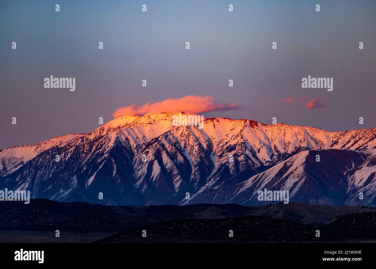 The pink light of the setting sun lights up a cloud over the three peaks of snow-capped Mount Nebo near Nephi, Utah, USA as seen from the west. Mount Stock Photo
