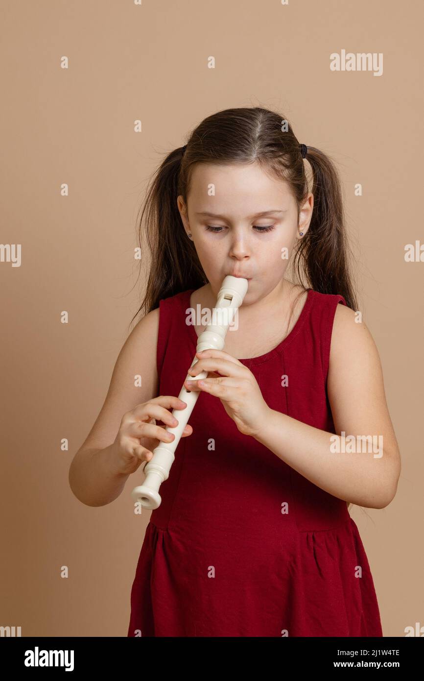 Girl in red dress play melody on flute with concentration, blowing air into duct, beige