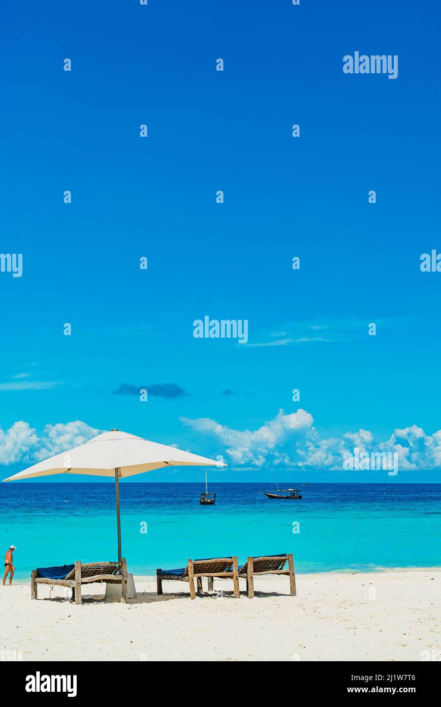 Wooden deck chairs on a sandy beach in front of ocean with boats, Zanzibar Tanzania Stock Photo