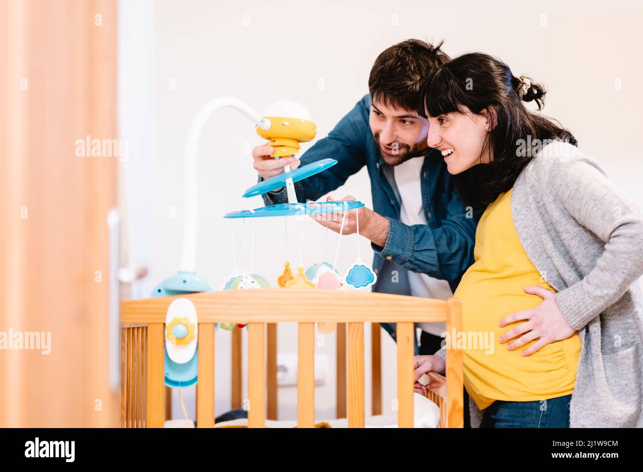 Happy husband and pregnant wife looking at toys hanging over cot placed