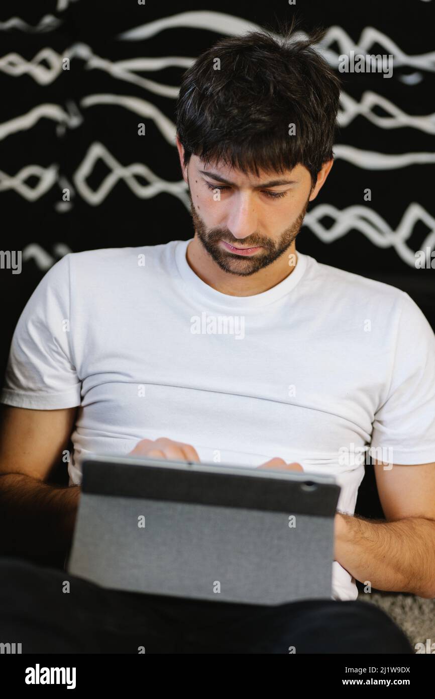 Concentrated young bearded ethnic male freelancer in white t shirt sitting on sofa and working remotely on tablet at home Stock Photo