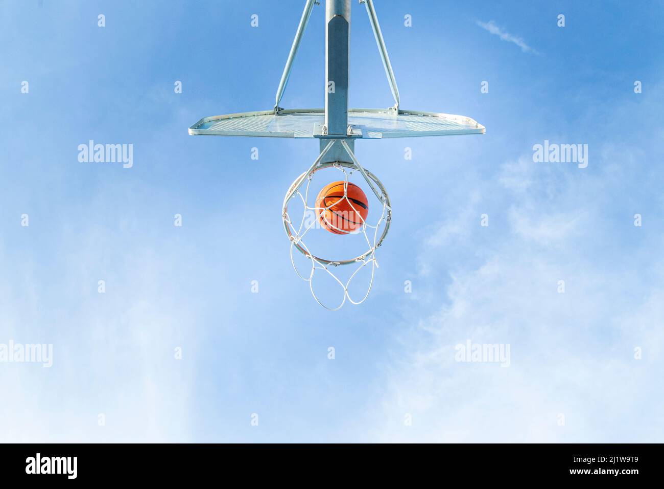 From below of orange basketball falling into round hoop with net placed against blue sky on sunny summer day in city Stock Photo