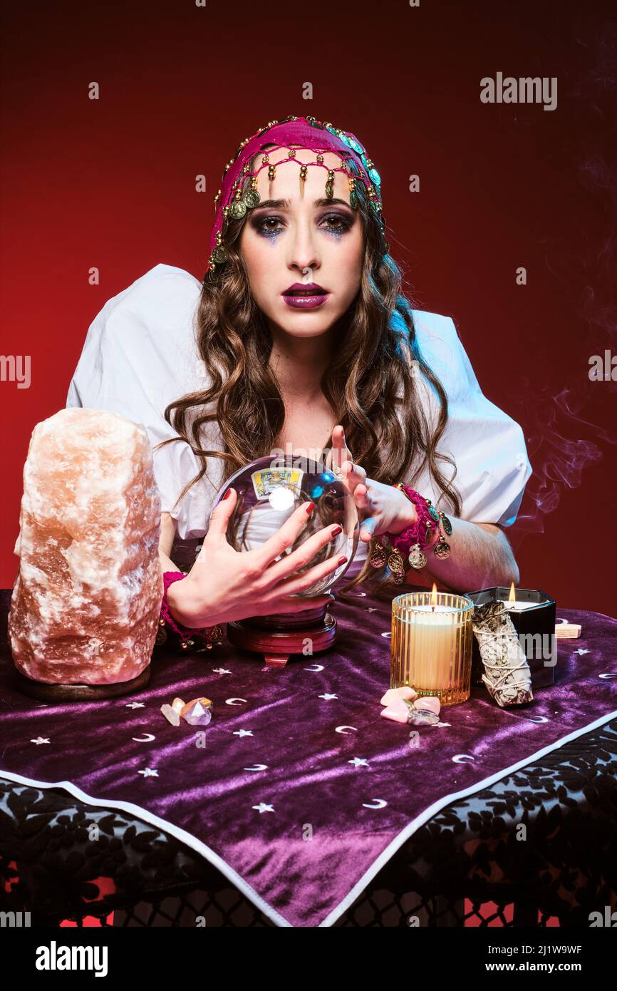 Charming female fortune teller with makeup guessing future at table with magic ball and burning candles near crystal Stock Photo