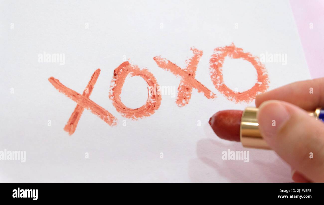 Closeup of hand holding a tube of red lipstick, with the symbol 'XOXO' written on a piece of paper. Stock Photo