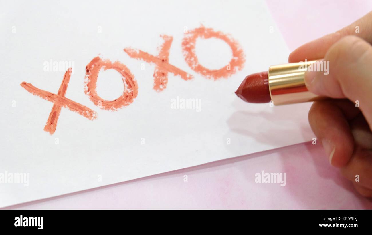 Hand holding a tube of red lipstick, with the symbol 'XOXO' written on a piece of paper. Stock Photo