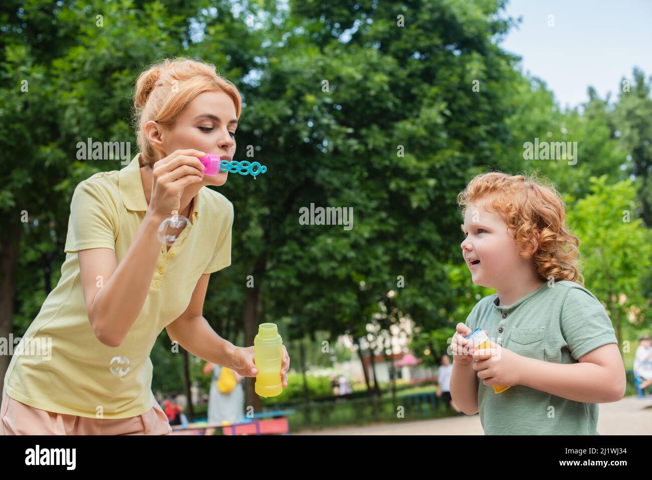 redhead woman blowing soap bubbles near excited son on playground Stock Photo