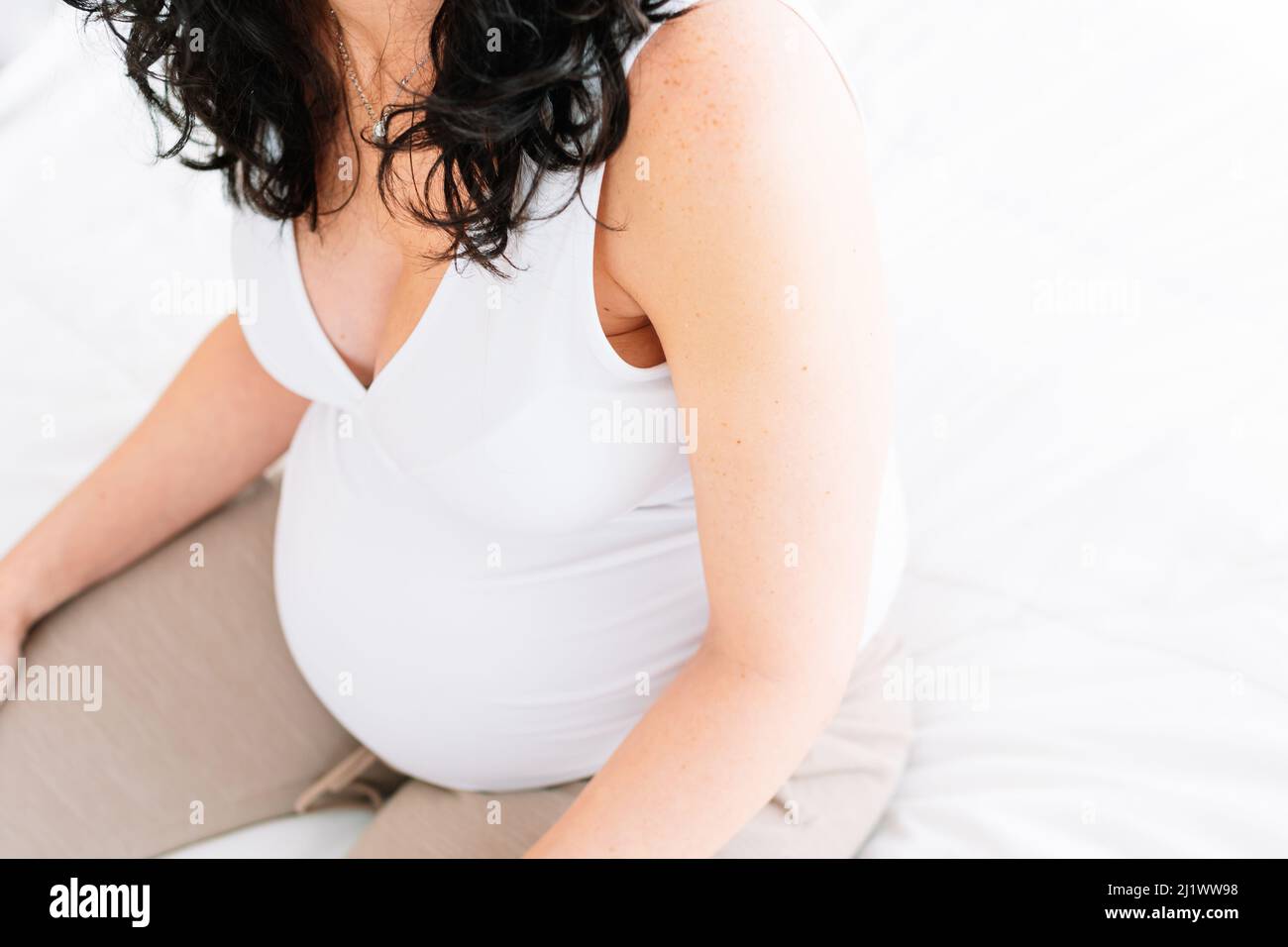 pregnant young woman with curly black hair is sitting on the white bed in her room. White background. copy space Stock Photo