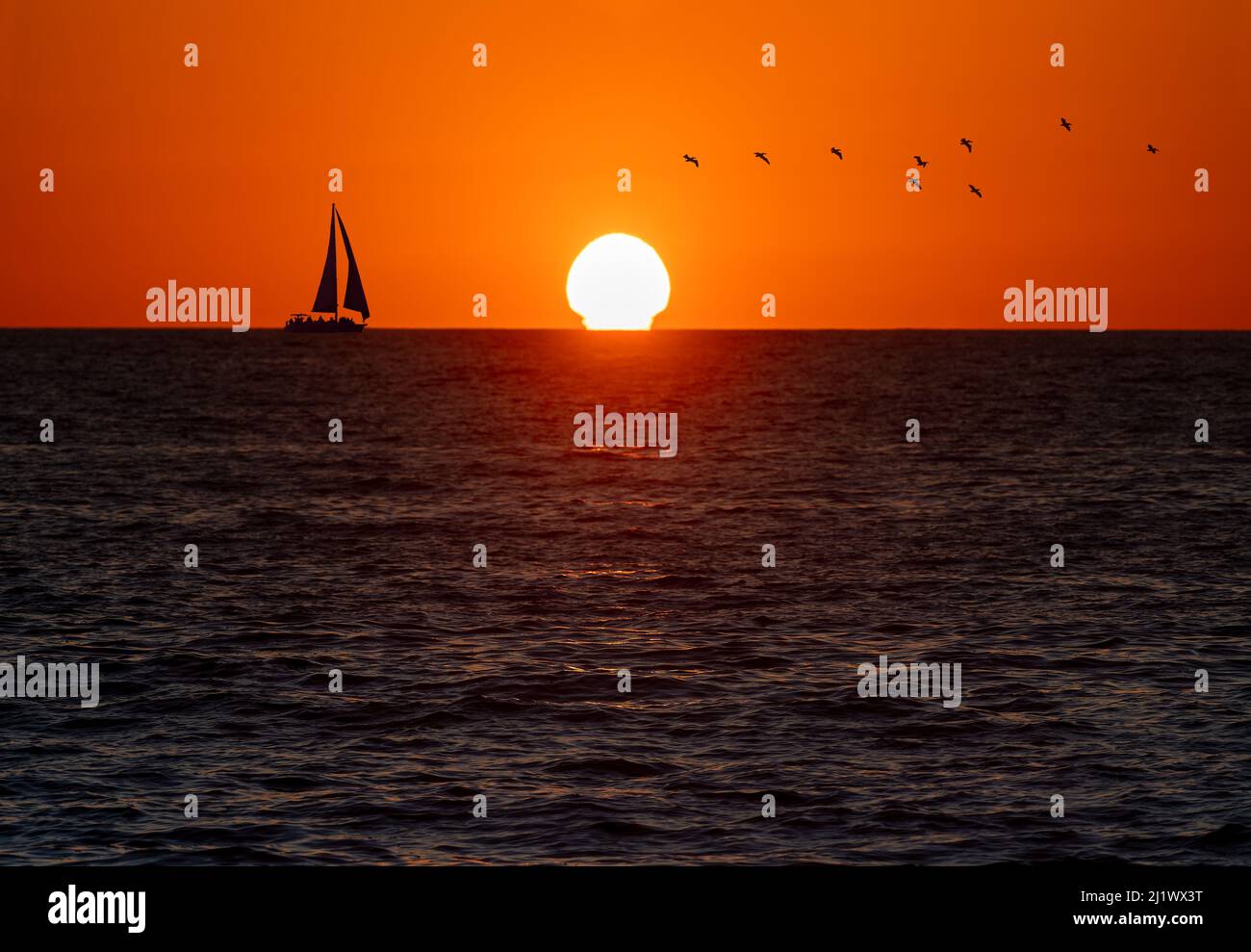 A Sailboat Sailing Along The Sea And Birds Flying Overhead In A Color Sunset Sky Stock Photo