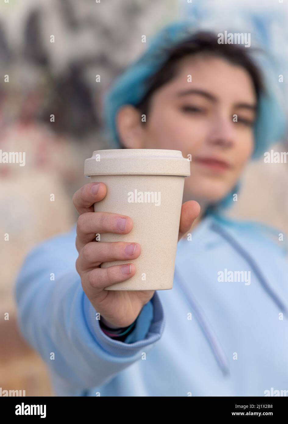 Teenagee in light blue oversize hoodie with coffee to go. Blue haired teen girl staying outdoors near graffiti wall. Cup mockup.  Hipster and adolesce Stock Photo