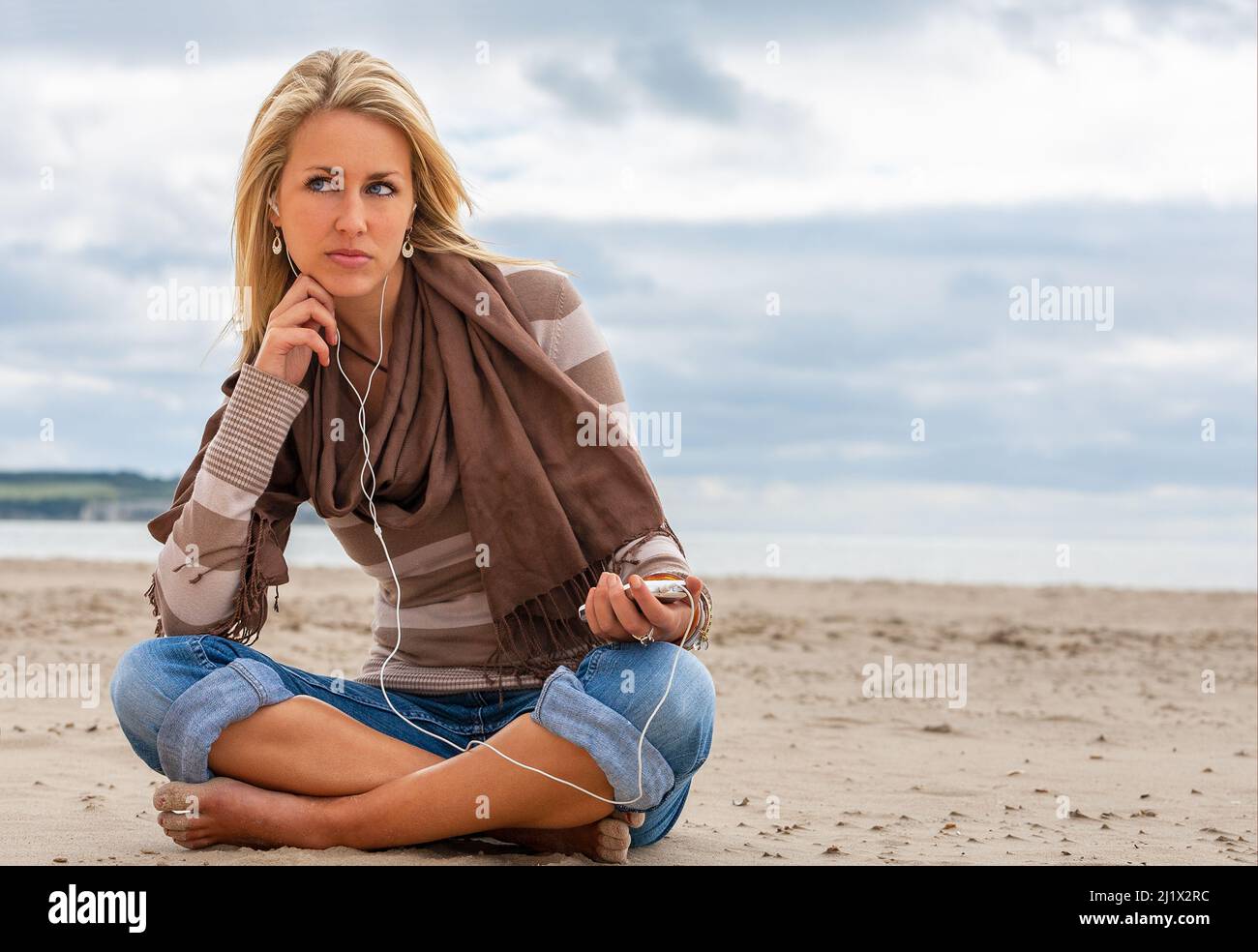 Beautiful thoughtful sads depressed young blond woman listening to music on a beach with her smartphone cell phone and headphones Stock Photo