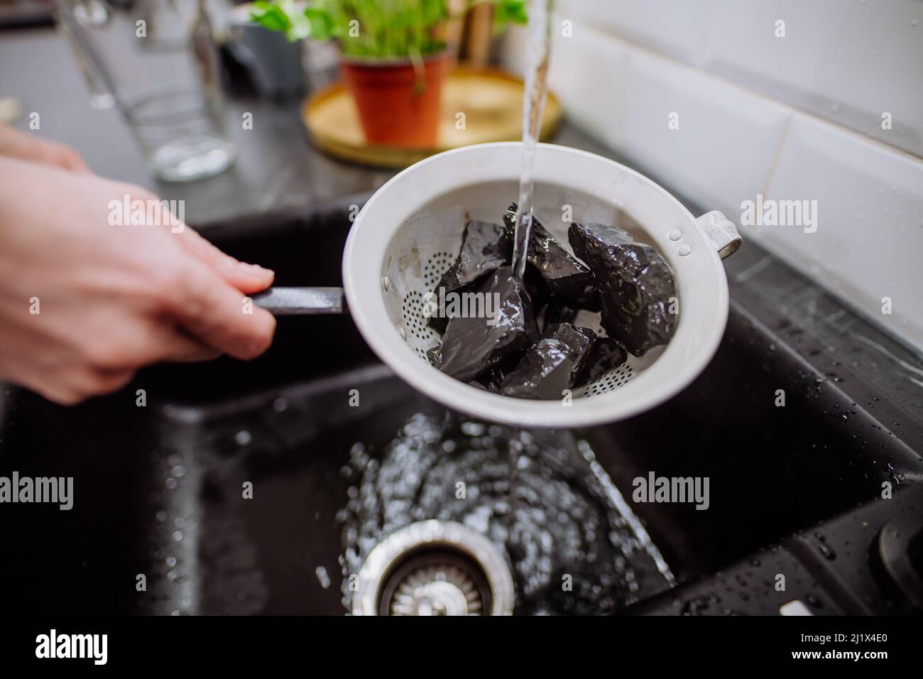 Woman cleaning shungite stones in sieve with pouring water in sink. Stock Photo