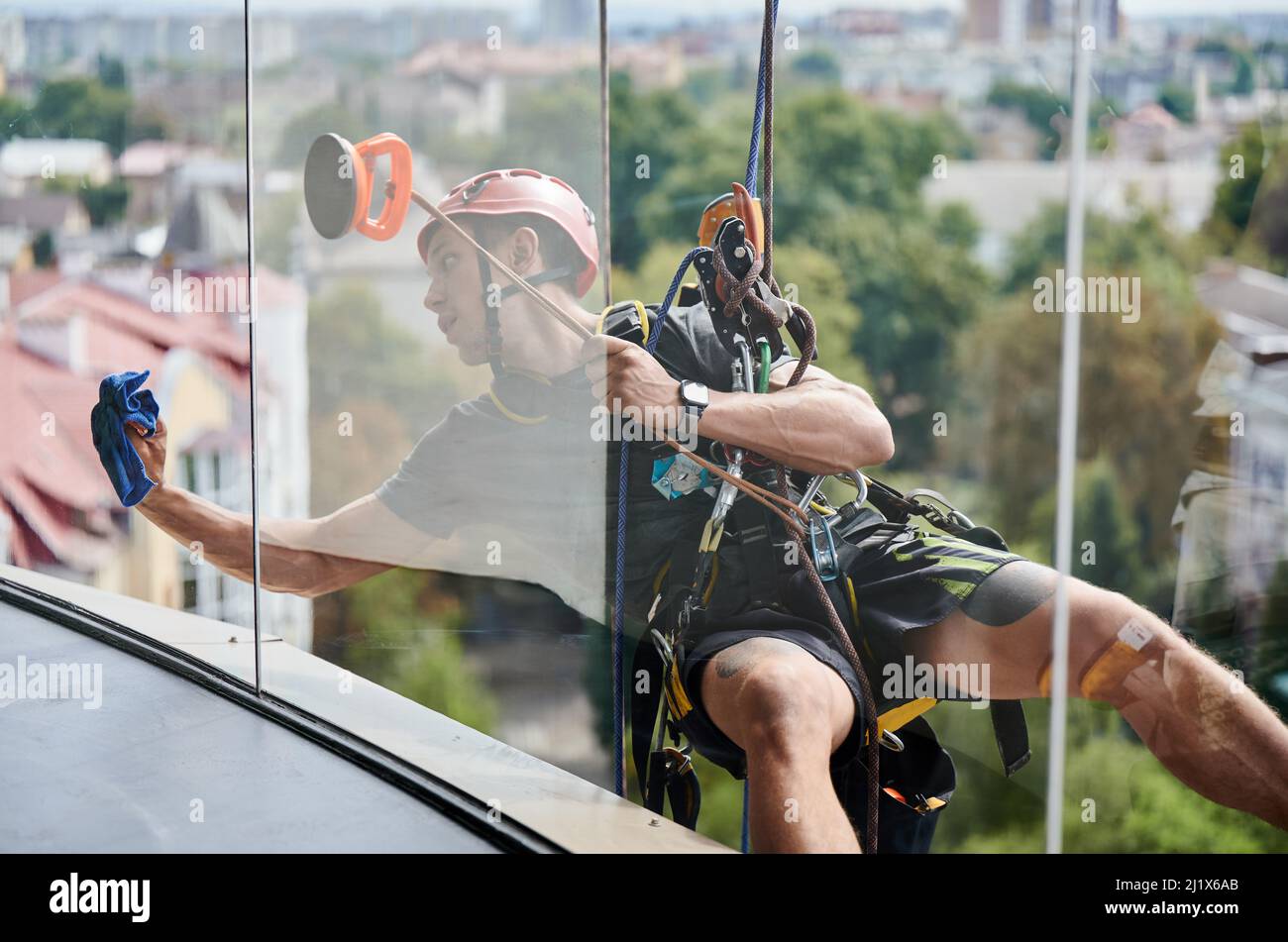 Industrial mountaineering cleaning service worker hanging on rope and wiping window. View from inside building. Cleaner using safety lifting equipment while cleaning glass of high-rise building. Stock Photo