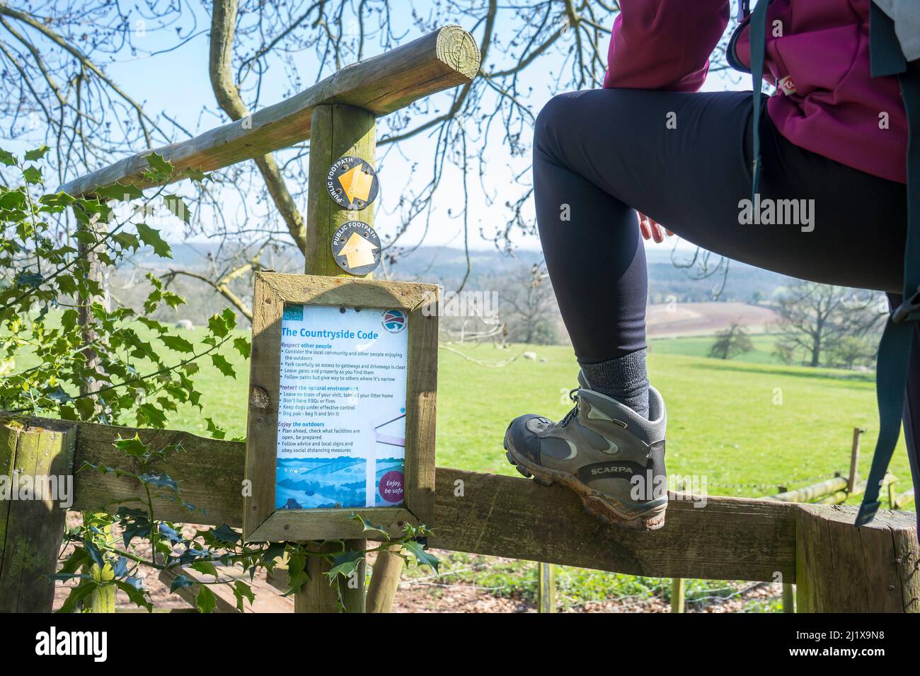 Leg close up of young, female hiker climbing over a wooden country stile with a countryside code sign fastened to it for hikers & ramblers to read. Stock Photo