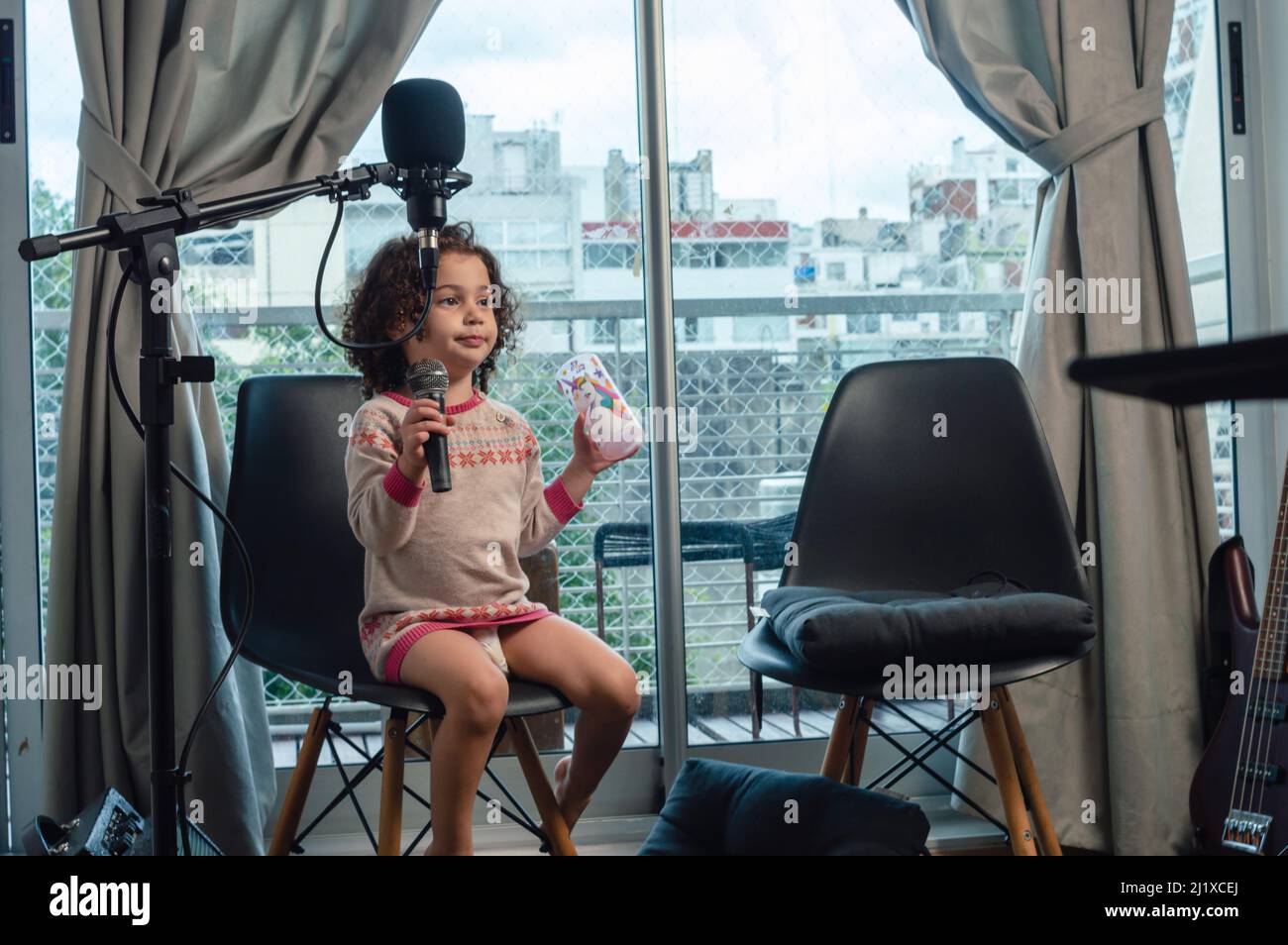 little venezuelan caucasian girl with curlers in diaper sitting in home music studio with microphone in hand, entertainment moment during work. Stock Photo