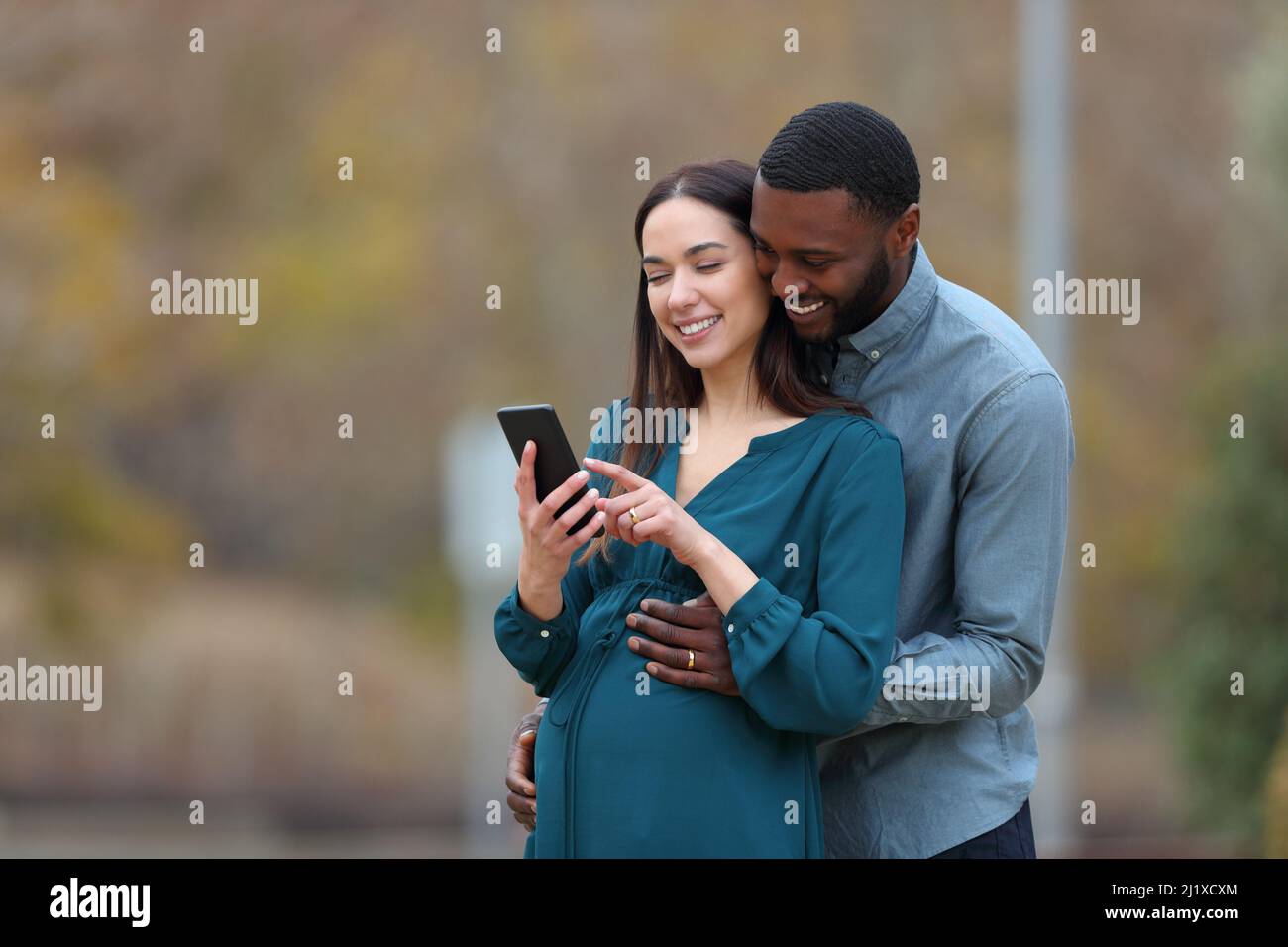 Interracial couple with a pregnant wife and her husband checking smart phone in a park Stock Photo