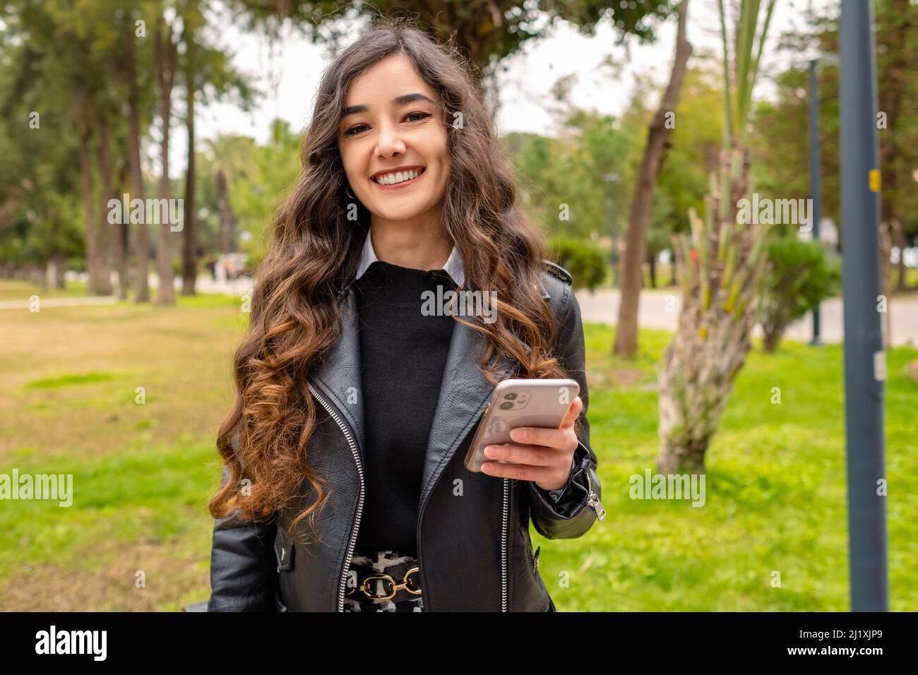 Smiling young lady, business woman posing for the camera and holding her phone, she is spending her time in an outdoor park and smiling, wavy hair and Stock Photo
