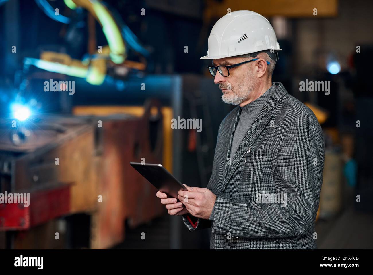 Serious elder engineer in work helmet using digital tablet to control the work on factory Stock Photo