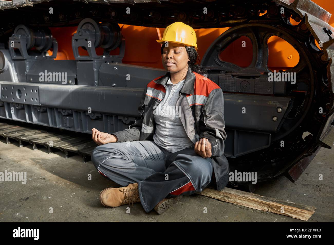 African female worker in uniform sitting in lotus position and meditating with tractor in the background Stock Photo