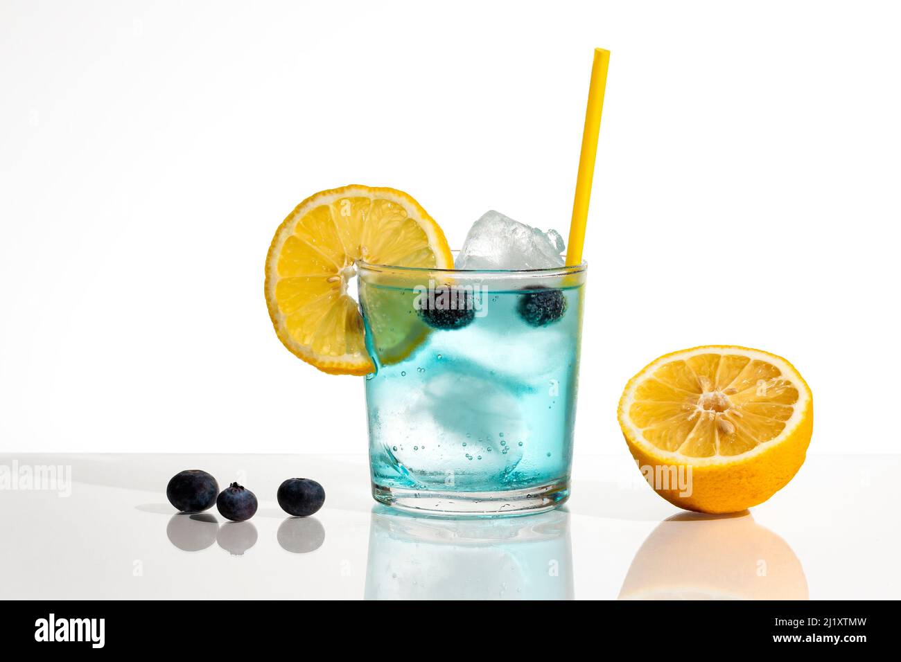 A glass of blue gin and tonic with ice, blueberries, a slice of lemon and a yellow straw. The background is white and the elements are reflected on th Stock Photo