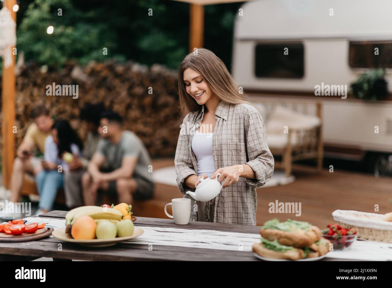 Happy millennial Caucasian woman pouring tea, making breakfast near RV, camping with her diverse friends outdoors Stock Photo