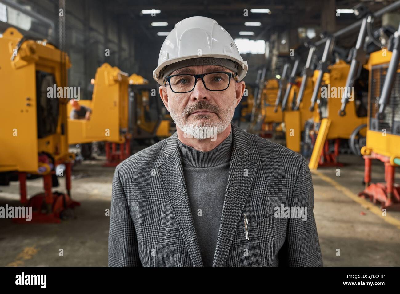 Portrait of senior engineer in work helmet looking at camera while standing in warehouse with new machines in the background Stock Photo