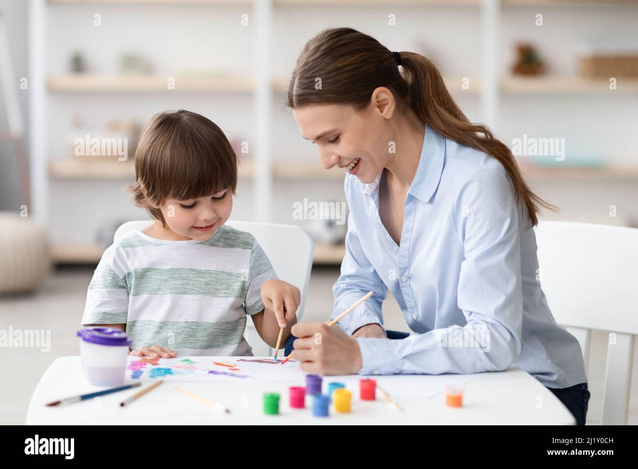 Cute little boy enjoying art classes, drawing picture with colorful paints, sitting at desk with teacher and laughing Stock Photo