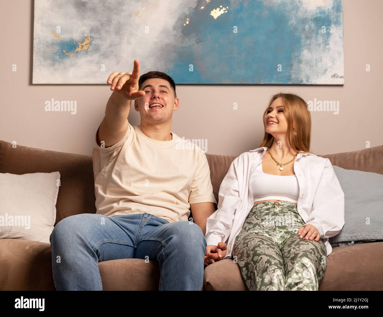 Minsk Belarus - 23 March 2022 Young couple sitting on couch, laughing and watching TV. Handsome man pointing at screen. Beautiful woman looking at husband or boyfriend. Happy spending time together . Stock Photo